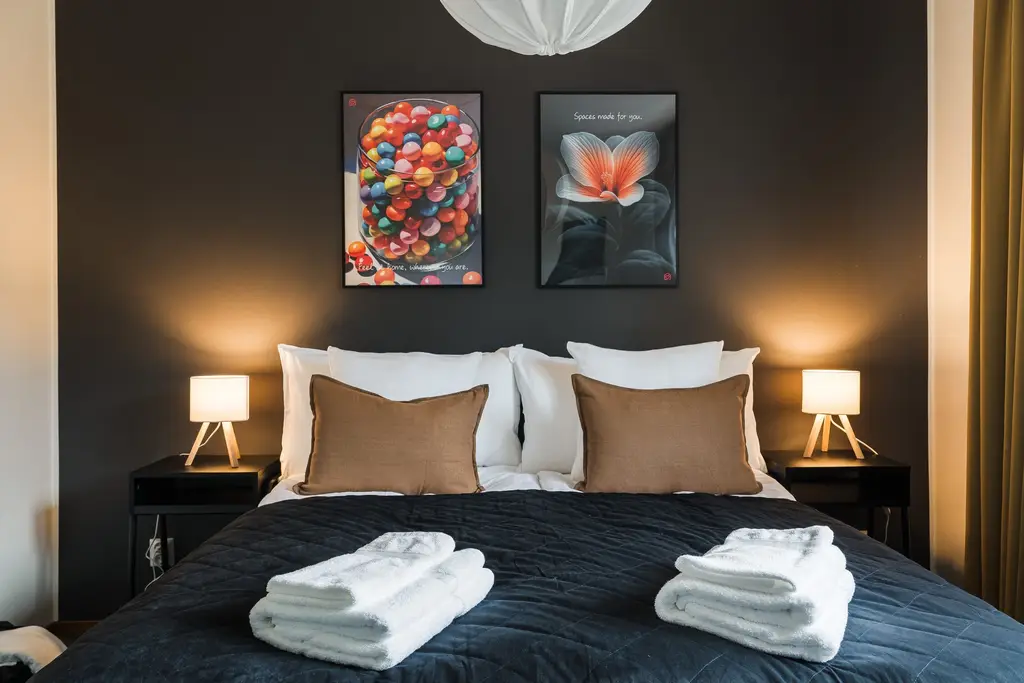 Sovrum med dubbelsäng mot en mörk vägg. Ovanför sängen hänger två inramade tryck med text: ett med kulörta glaskulor och orden Feel at home, wherever you are, och ett med en blomma och orden Spaces made for you. Bruna och vita kuddar, mörkgrått överkast, vikta handdukar på fotändan och sängbord med tända bordslampor på vardera sida.
