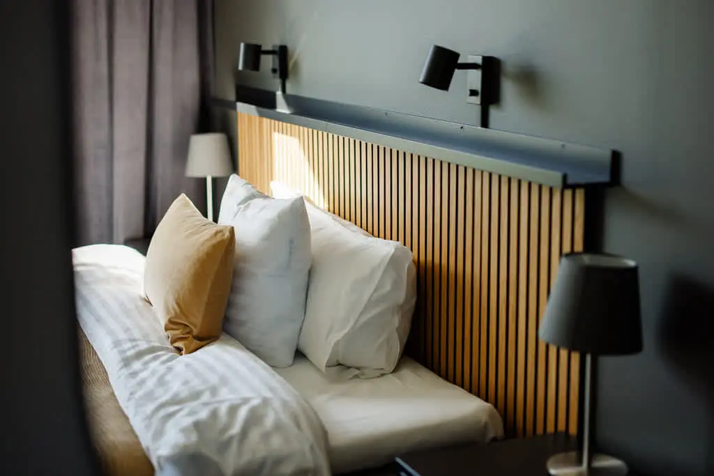 Säng med vita och sandfärgade kuddar mot en ribbad sänggavel i ljust trä, två svarta läslampor monterade på väggen ovanför, ett gråbeige draperi till vänster och en bordslampa på nattduksbordet.