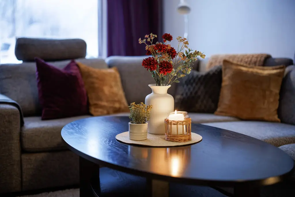 Vardagsrum med grå tygsoffa, kuddar i vinrött och senapsgult, ett ovalt mörkt soffbord med en vas med röda och torkade blommor, en liten kruka och ett tänt ljus i en korglykta. Fönster med draperi bakom.