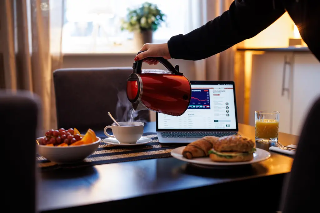En person häller upp hett vatten i en kopp vid ett matbord där en bärbar dator står uppfälld bredvid frukost med frukt, croissant och juice.